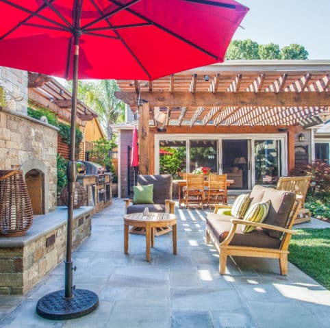 Patio with wooden furniture under a red umbrella and pergola, next to a stone wall.